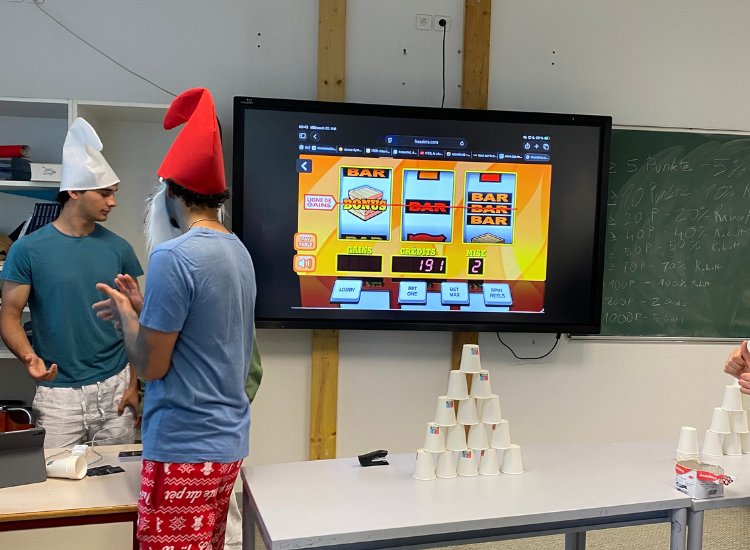Schüler neben einem Bildschirm mit Glücksspiel, davor eine Becherpyramide