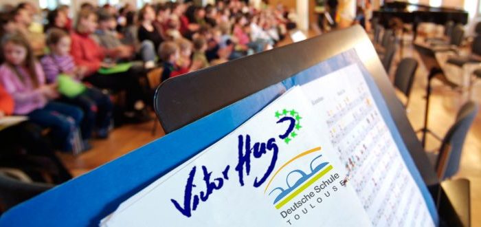 Notenblatt mit den Logos der Deutschen Schule Toulouse und Lycée Victor Hugo, im Hintergrund Kinder im Publikum
