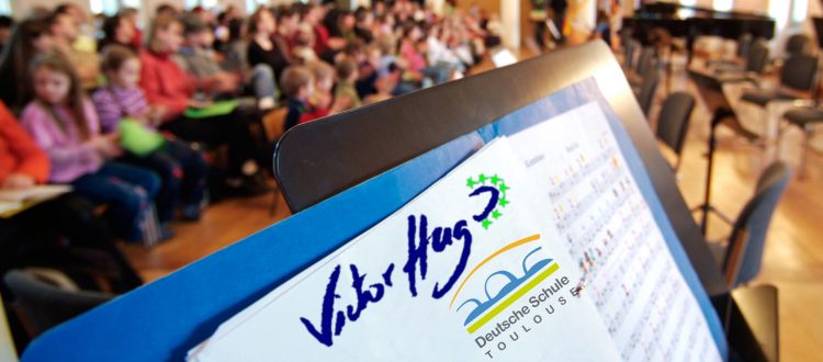 Notenblatt mit den Logos der Deutschen Schule Toulouse und Lycée Victor Hugo, im Hintergrund Kinder im Publikum